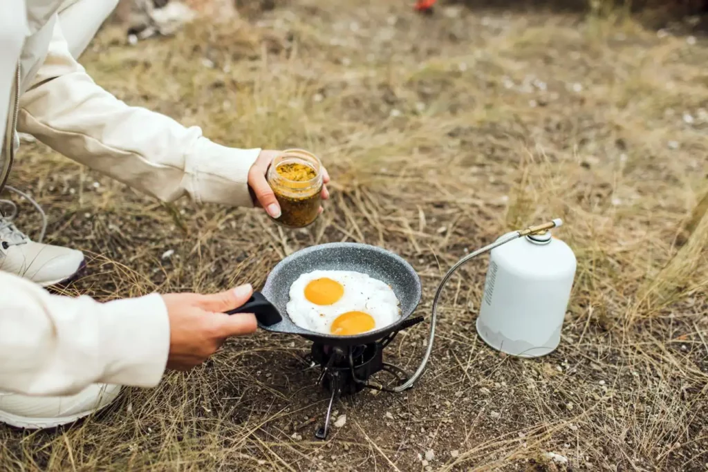 Young man cooking fried eggs outdoors with camping gas stove